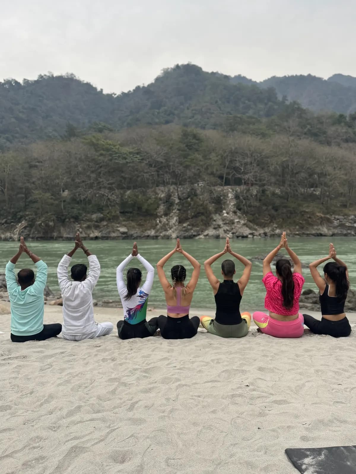 Retreat guests meditating with arms raised on the Ganga riverbank in Rishikesh